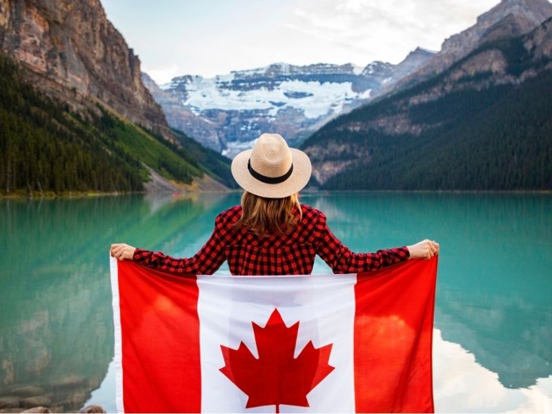 Female Standing With A Red and White Canadian Flag Around Her Looking At the Beautiful Mountains And Lakes In The Rocky Mountains in Canada.