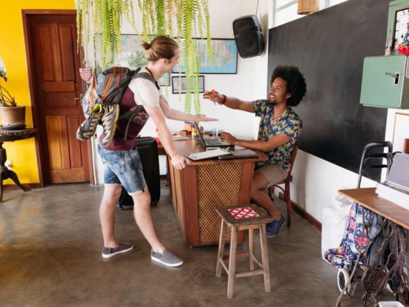Backpacker Receiving Key From Receptionist At Hostel Reception. Before You Receive The Key To Your Dorm Room In The Hostel From The Receptionist Make Sure You Do Your Research That It Offers The Services You Require.
