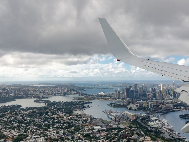 Looking Out A Plane Window While Landing Over Sydney Harbour With The Harbour Bridge And The Sydney Opera House Standing Out Amongst The Buildings.