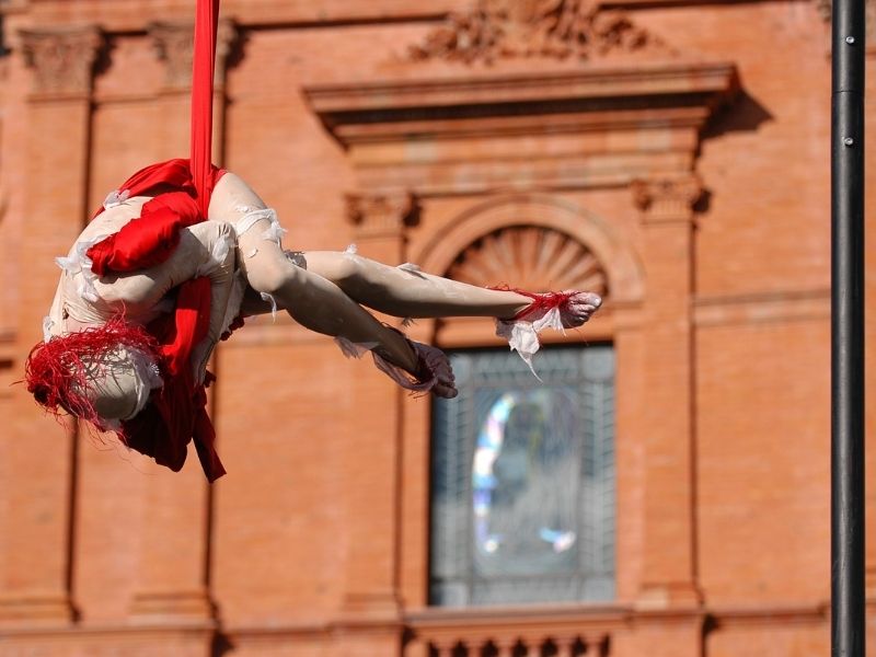 A Female Acrobat Hanging By Red Material And Twisting Around As She Busks On The Street.