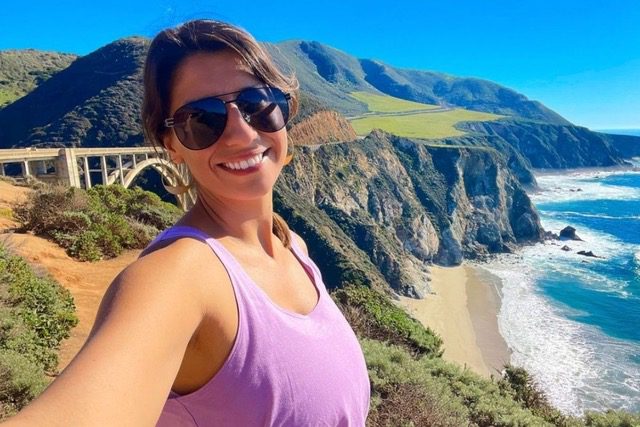 Female On The Big Sur Road Trip In America. Taking A Selfie With The Road Behind.