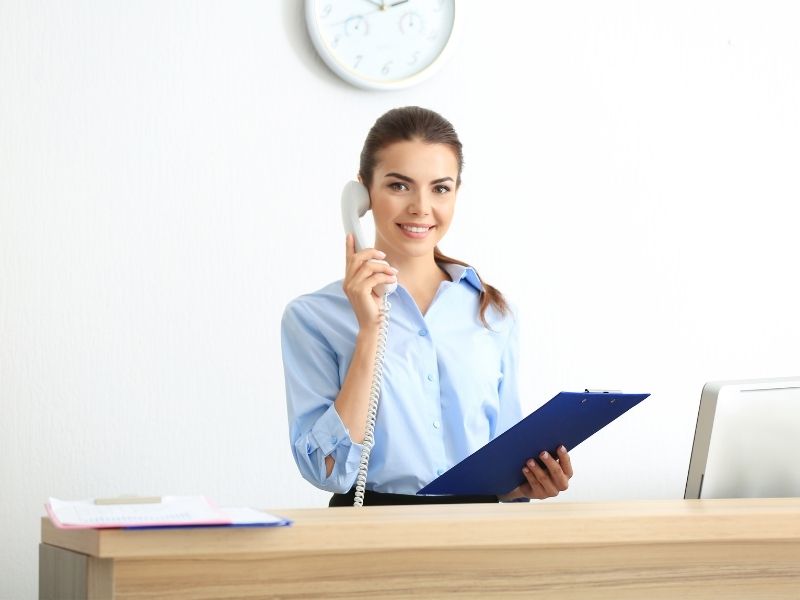 Female Answering A Phone Behind A Desk In An Office As The Receptionist. This Is A Great Working Holiday Job.