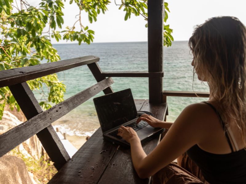 Female in swimwear sitting at a table with a laptop on a deck overlooking the ocean.