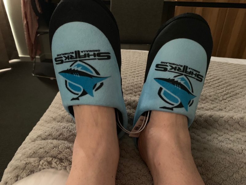 Cronulla Sharks slippers on feet on a bed.